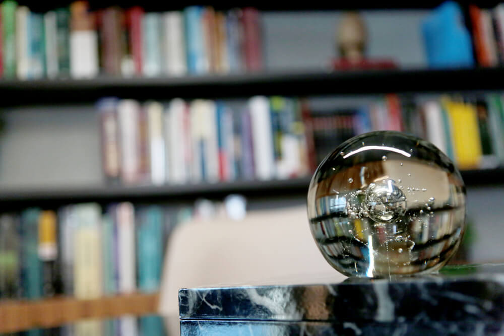 Em primeiro plano um globo de vidro aparece sobre uma mesa, atrás, desfocada, uma grande estante com livros